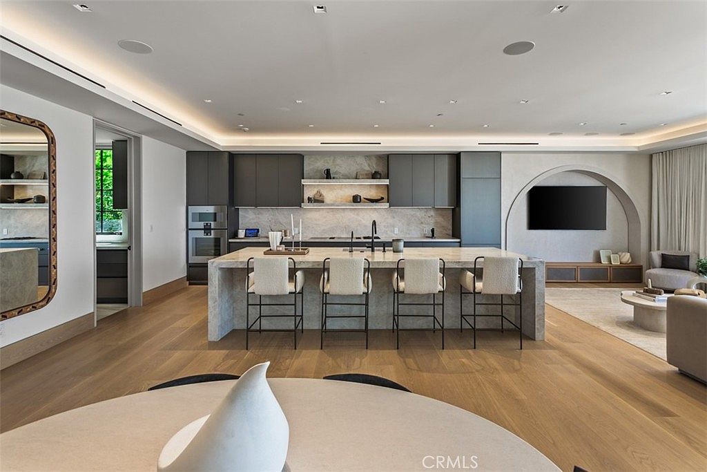 This sophisticated, open-concept kitchen features a large stone-topped island with four modern bar stools, serving as the centerpiece of the room. The space is defined by sleek, dark grey cabinetry, integrated high-end appliances, and warm wood flooring that flows into an adjacent living area with an arched wall feature. Recessed lighting and a minimalist aesthetic create a bright, airy, and luxurious atmosphere perfect for contemporary living.