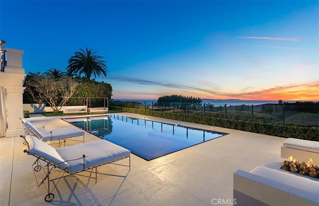 This stunning outdoor space features a sleek, rectangular infinity-edge pool that seamlessly blends into the horizon, overlooking a breathtaking sunset over the ocean. The expansive stone patio is furnished with modern lounge chairs and a cozy fire pit area, creating an ideal setting for luxury relaxation and entertaining. The composition captures a serene, high-end atmosphere with a clear view of the coastal landscape.