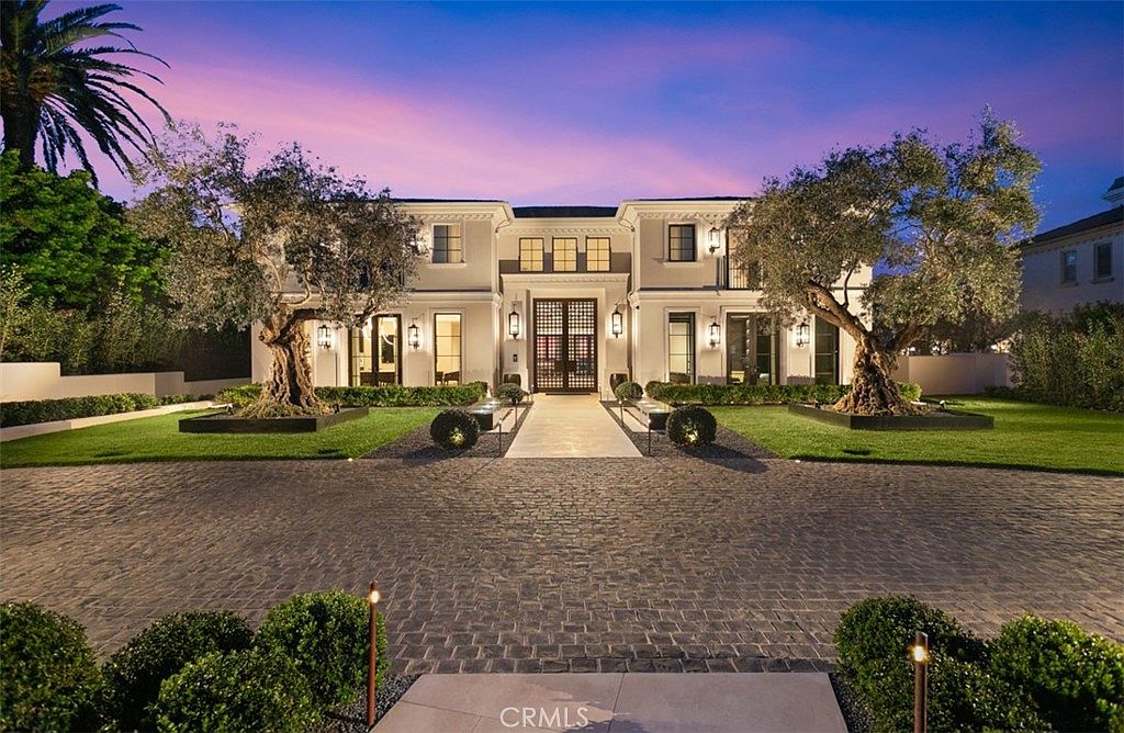 This stunning luxury estate features a symmetrical, grand facade with a prominent central entryway and elegant architectural detailing. The property is framed by two mature, sculptural olive trees and a meticulously manicured lawn, all set against a dramatic twilight sky. A cobblestone driveway leads directly to the impressive front door, creating a sophisticated and welcoming first impression.