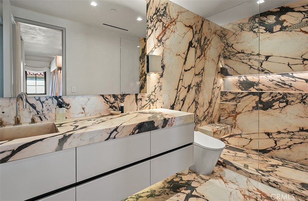 This luxurious primary bathroom features dramatic, high-contrast marble slab walls and flooring that create a sophisticated, high-end aesthetic. The space includes a sleek, floating white vanity with a matching marble countertop and a seamless glass-enclosed walk-in shower. The modern design is complemented by recessed lighting and a minimalist toilet, offering a clean and opulent atmosphere.