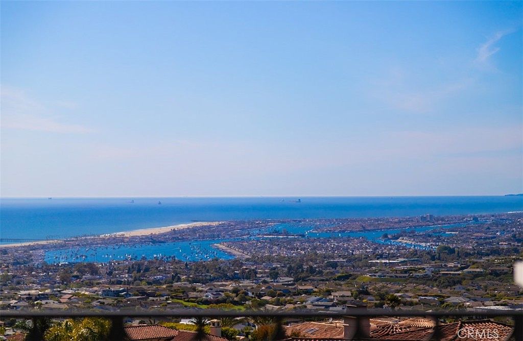 This image is captured from an elevated balcony, offering a breathtaking, expansive panoramic view of a coastal harbor and the ocean beyond. The foreground features a dark railing that frames the scene, looking down over a dense residential neighborhood nestled into the hillside. The clear blue sky and sparkling water create a serene and luxurious atmosphere, highlighting the property's prime location and scenic outlook.