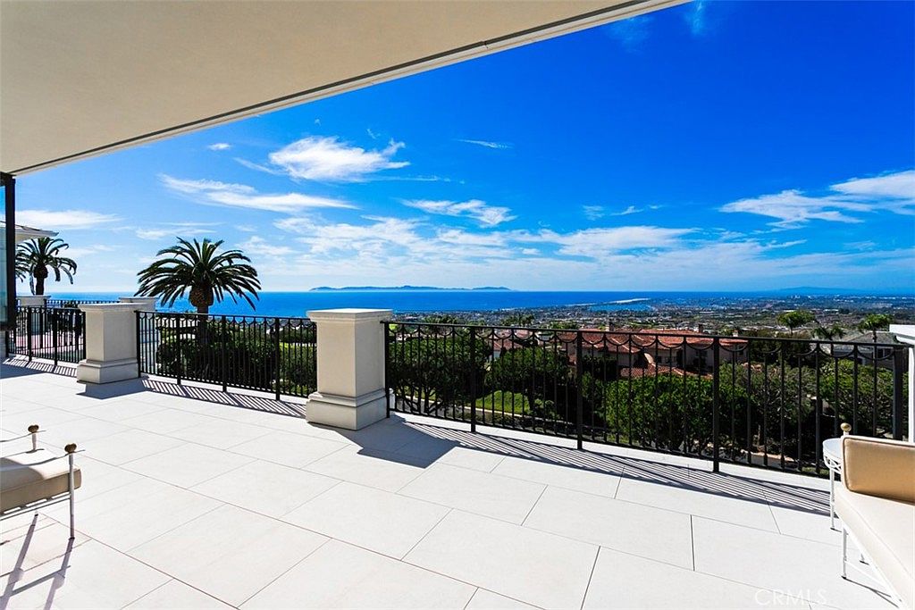 This expansive, high-end balcony offers a breathtaking panoramic view of the coastline, ocean, and distant islands. The space features sleek, light-colored stone flooring and a classic black wrought-iron railing, creating a sophisticated and airy atmosphere. The perspective from the covered terrace emphasizes the seamless transition between luxury indoor living and the stunning natural landscape.