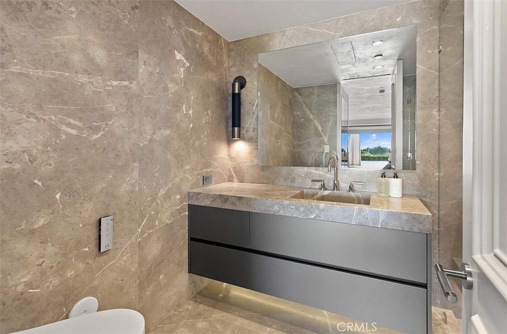 This luxurious primary bathroom features floor-to-ceiling marble tiling that creates a sophisticated and cohesive aesthetic. The floating vanity is topped with a matching marble slab and a sleek integrated sink, complemented by a large, frameless mirror and modern wall-mounted lighting. The perspective captures the elegant vanity area, highlighting the high-end finishes and the clean, contemporary design.