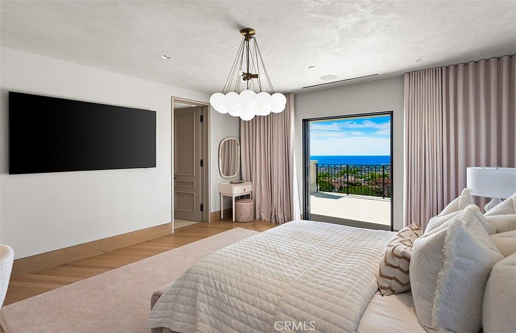 This luxurious primary bedroom features a neutral, monochromatic color palette with soft textures and a sophisticated, modern aesthetic. A large, wall-mounted television sits opposite a plush bed, while a sleek vanity area and floor-to-ceiling curtains frame a sliding glass door that opens to a balcony with stunning ocean views. The room is illuminated by a unique, multi-globe chandelier, creating an elegant and serene atmosphere.