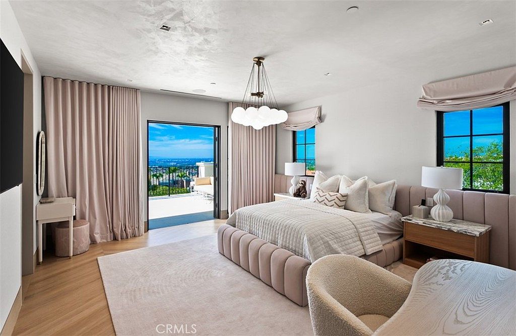 This luxurious primary bedroom features a soft, neutral color palette with plush, upholstered furniture and elegant drapery. A large glass door opens onto a private balcony, offering a stunning panoramic view of the landscape beyond. The room is anchored by a modern cloud-like chandelier and a sophisticated, contemporary design aesthetic.