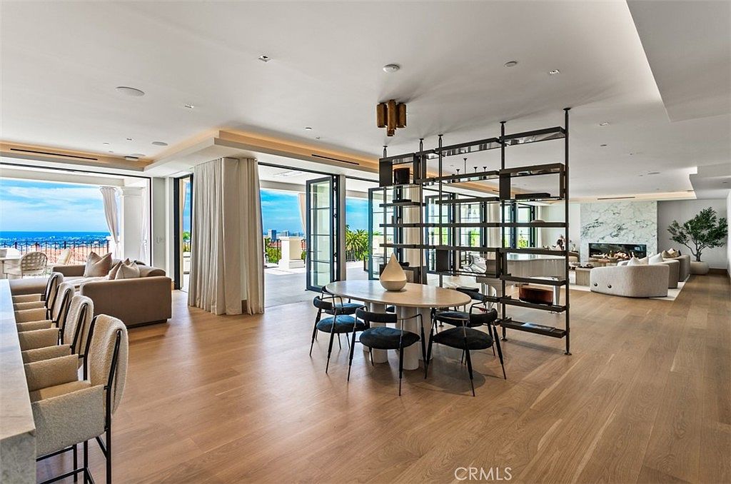 This expansive, open-concept living and dining area features light hardwood flooring, high ceilings with recessed lighting, and a sophisticated metal-framed room divider. The space seamlessly transitions to an outdoor balcony through large glass doors, offering a stunning panoramic ocean view. The interior design is modern and minimalist, characterized by neutral tones, plush seating, and a marble-clad fireplace that anchors the living space.
