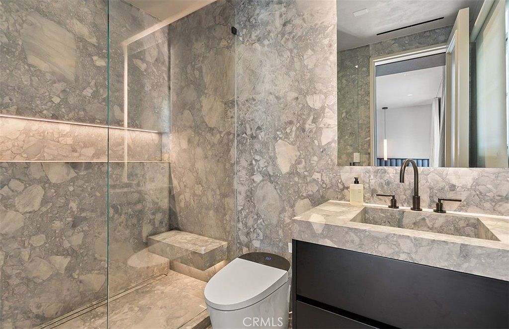 This luxurious, modern bathroom features striking grey-veined marble slab walls and countertops that create a cohesive, high-end aesthetic. The space includes a sleek, wall-mounted toilet, a floating vanity with dark cabinetry, and a spacious walk-in shower with a built-in marble bench. The perspective is a straight-on shot that highlights the sophisticated materials and clean, minimalist design.
