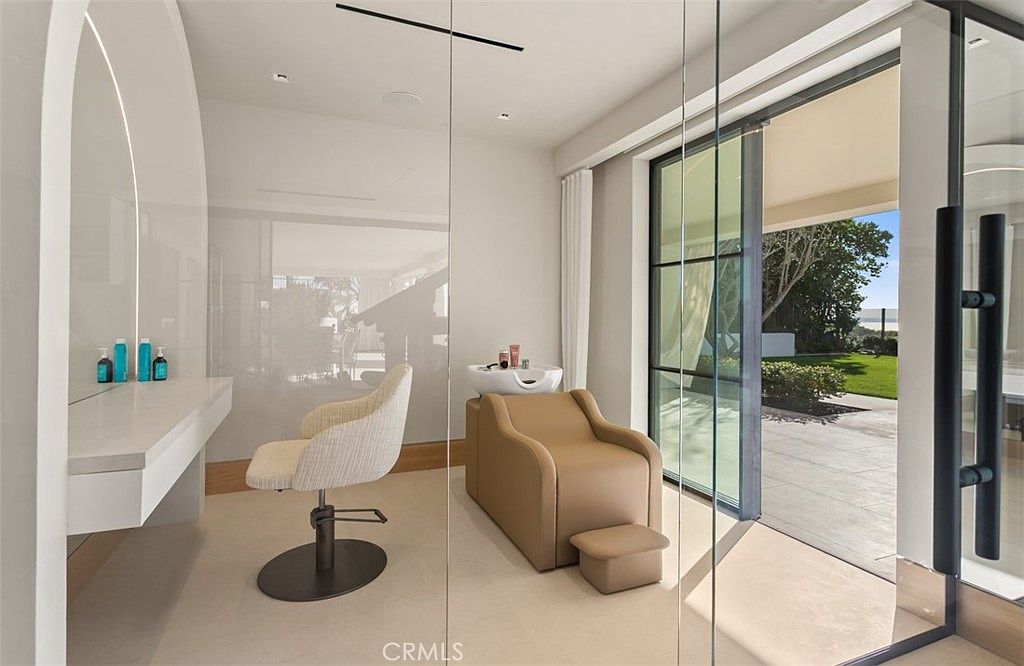 This image showcases a luxurious, private salon or spa room located within a residence, featuring a professional-grade hair washing station and a dedicated styling chair. The space is designed with a minimalist, modern aesthetic, utilizing neutral tones, sleek glass partitions, and large sliding doors that open to a serene outdoor garden view. The room offers a high-end, tranquil atmosphere perfect for personal grooming and relaxation.