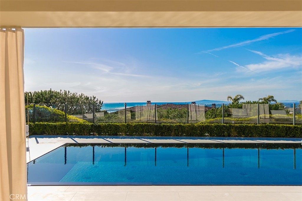 This stunning outdoor space features a sleek, rectangular infinity-edge pool that seamlessly blends into a breathtaking panoramic view of the ocean and coastline. The foreground includes a clean, light-toned patio area framed by a manicured hedge and a modern glass railing, creating an unobstructed connection to the horizon. The perspective is captured from a covered patio, emphasizing a luxurious, serene, and high-end coastal living experience.