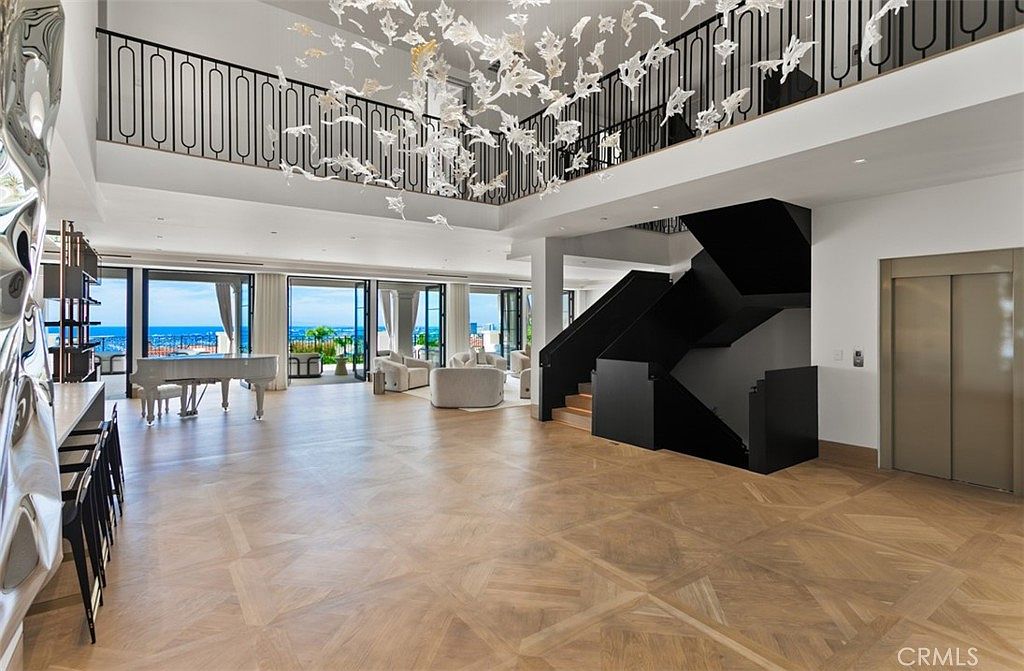 This expansive, open-concept living area features a dramatic double-height ceiling adorned with a striking, sculptural chandelier. The space is anchored by light-toned herringbone wood flooring, a modern black staircase, and a sleek elevator, all framed by floor-to-ceiling glass doors that reveal a stunning ocean view. The room exudes a sophisticated, contemporary luxury aesthetic, perfect for grand entertaining.