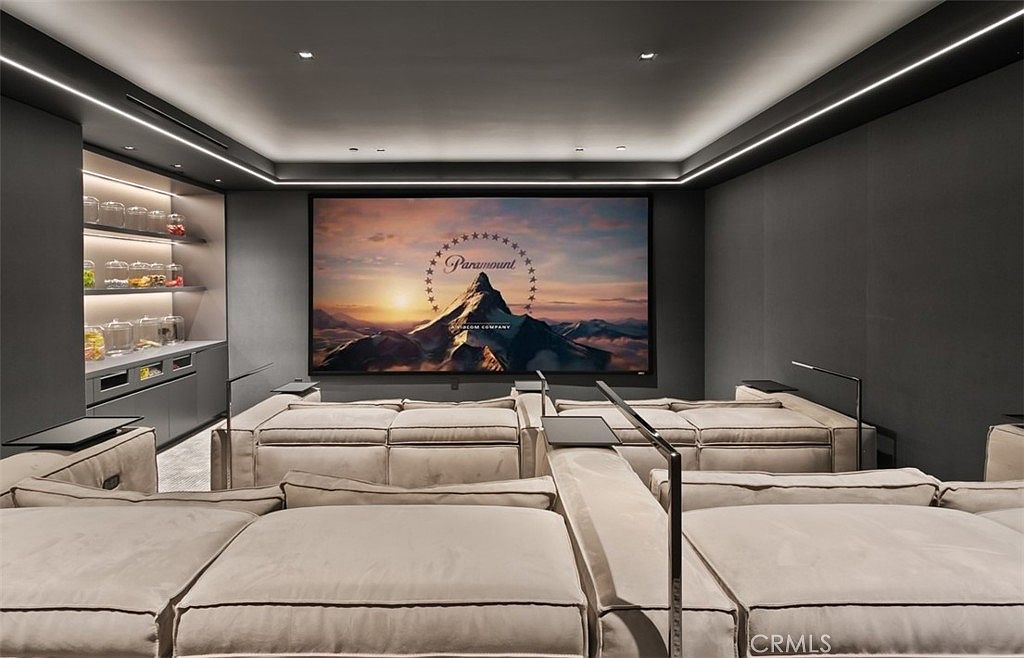 This image showcases a luxurious, modern home theater designed for an immersive cinematic experience. The room features tiered, plush light-colored seating, a large projection screen displaying the Paramount logo, and integrated ambient cove lighting that creates a sophisticated atmosphere. To the side, a built-in snack bar with illuminated shelving adds a functional and high-end touch to the entertainment space.
