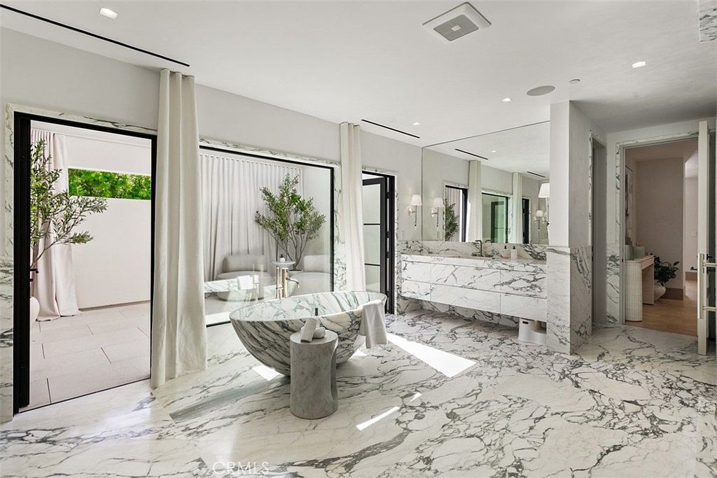 This luxurious primary bathroom features stunning floor-to-ceiling marble veining throughout, centered around a dramatic, sculptural stone soaking tub. The space is bright and airy, with large glass doors leading to a private outdoor area and a sleek, expansive vanity with a large mirror. The overall aesthetic is one of high-end, contemporary elegance and serene sophistication.