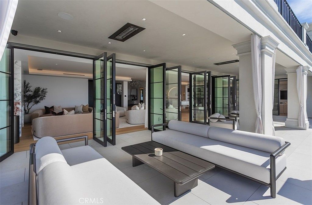 This luxurious covered patio features a seamless indoor-outdoor transition through expansive, multi-panel folding glass doors that open into a sophisticated living area. The space is furnished with modern, minimalist outdoor seating, including a sleek sofa and a low-profile coffee table, all set against a backdrop of elegant columns and neutral stone flooring. The overall aesthetic is one of high-end, contemporary comfort, perfect for refined outdoor entertaining.