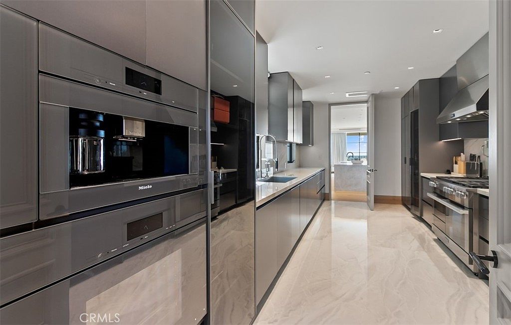 This sleek, modern galley kitchen features high-end Miele appliances, including a built-in coffee system and a professional-grade range. The space is defined by its minimalist dark grey cabinetry, polished light marble flooring, and clean, linear design. A perspective shot looking down the length of the kitchen highlights the seamless integration of luxury appliances and the sophisticated, contemporary aesthetic.