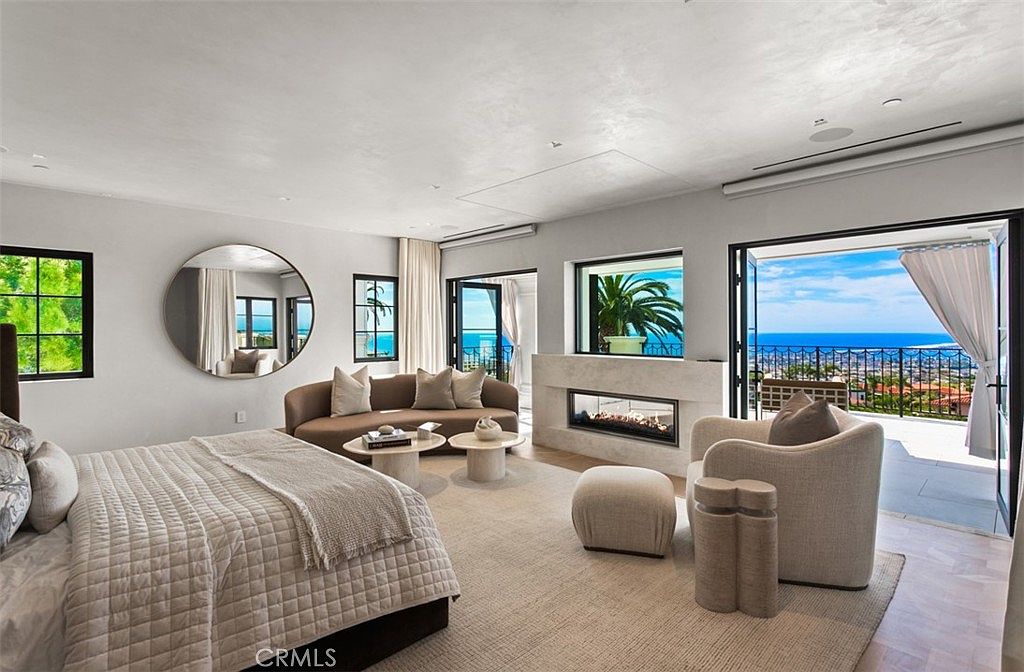 This luxurious primary bedroom features a sophisticated, neutral color palette and a seamless transition to a private balcony with stunning ocean views. The room is anchored by a plush bed, a curved sofa, and a modern linear fireplace, all set against a backdrop of soft, textured walls and high-end finishes. The open-concept design and large glass doors create an airy, serene atmosphere that emphasizes the home's coastal location.