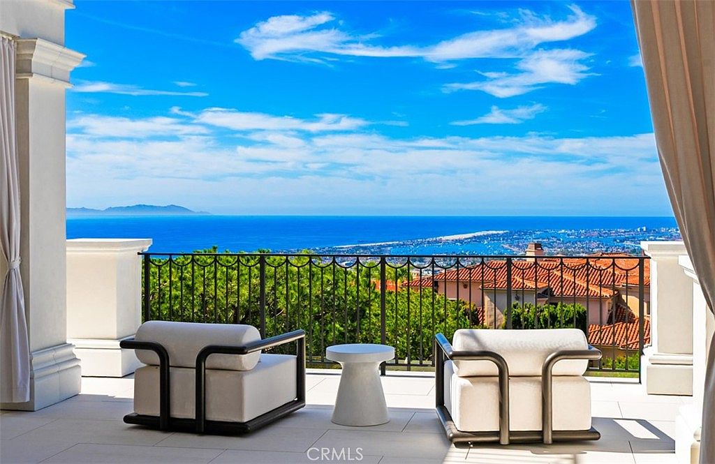 This elegant, covered balcony offers a breathtaking panoramic view of the ocean and coastline, framed by white architectural columns and soft, flowing curtains. The space is furnished with two modern, plush lounge chairs and a minimalist side table, creating a serene and luxurious outdoor retreat. The bright, sunny atmosphere and expansive horizon emphasize the property's prime location and high-end lifestyle appeal.