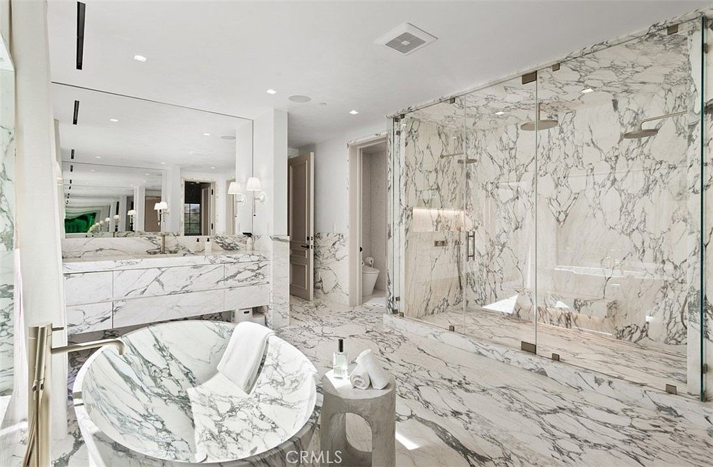 This luxurious primary bathroom features extensive use of white marble with dramatic grey veining throughout the walls, floors, and vanity. The space includes a large, freestanding soaking tub, a spacious walk-in glass shower, and a double vanity with a large mirror, creating an elegant and spa-like atmosphere. The bright, high-end design is complemented by modern fixtures and clean, minimalist lines.