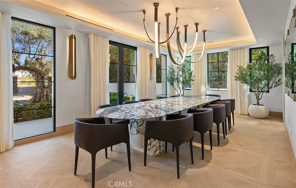 This elegant dining room features a long, oval marble table surrounded by sleek, dark modern chairs, creating a sophisticated atmosphere. A striking, multi-armed contemporary chandelier serves as the focal point above the table, while large floor-to-ceiling windows provide ample natural light and views of the outdoor greenery. The room is finished with light-toned herringbone wood flooring and minimalist wall sconces, offering a clean, luxurious aesthetic.