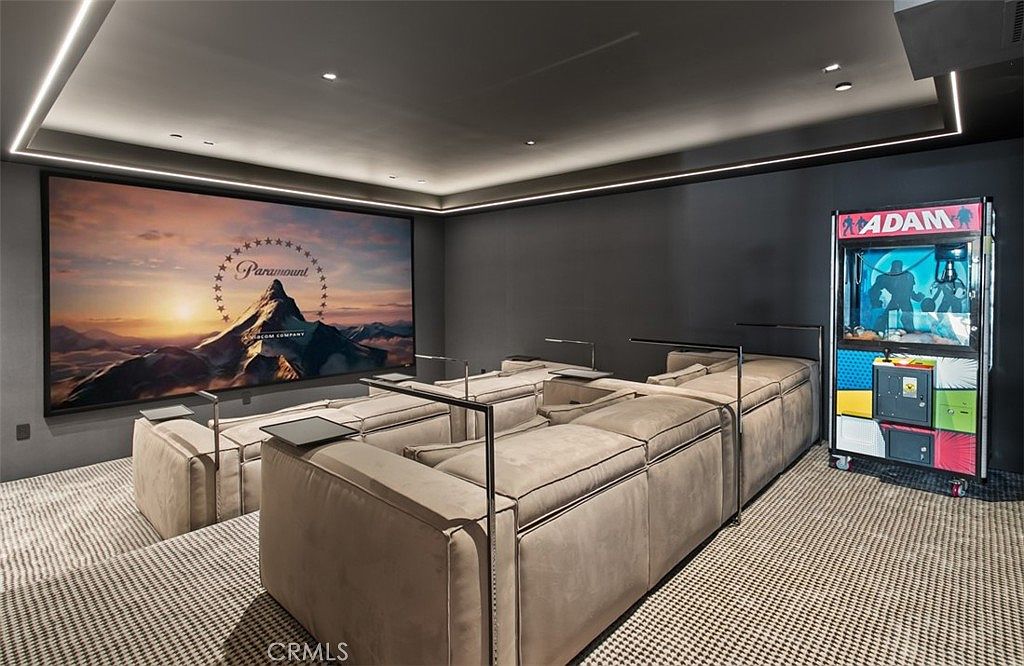 This image showcases a luxurious, modern home theater room designed for an immersive cinematic experience. The space features plush, tiered sectional seating, a massive wall-mounted projection screen displaying the Paramount logo, and a playful claw machine in the corner, all set against a dark, moody color palette with recessed LED lighting. The perspective is from the back of the room, emphasizing the comfortable, high-end entertainment environment.