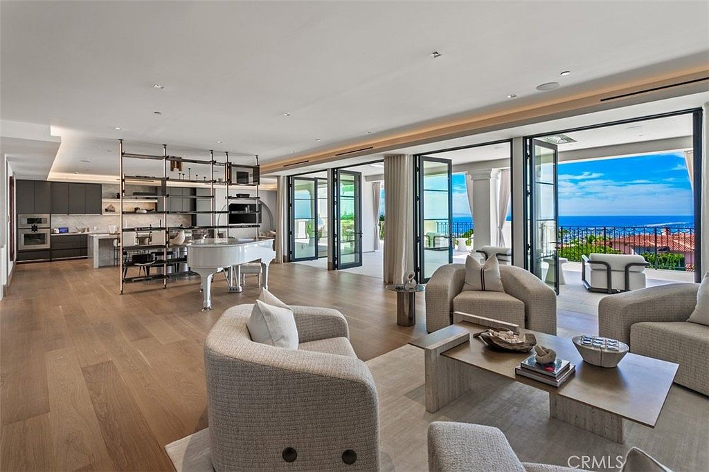 This expansive, open-concept living area features light hardwood flooring, a grand white piano, and a sophisticated seating arrangement centered around a modern coffee table. Large floor-to-ceiling glass doors open onto a balcony with stunning ocean views, seamlessly blending indoor and outdoor living. The space is characterized by a neutral color palette, contemporary furnishings, and a sleek, minimalist aesthetic that emphasizes luxury and natural light.