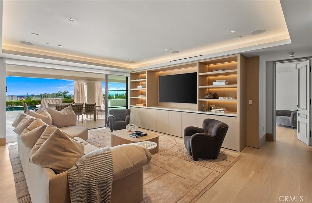 This sophisticated living room features a seamless indoor-outdoor flow, with large sliding glass doors opening onto a sun-drenched patio and pool area. The space is anchored by a custom light-wood built-in media wall with integrated shelving and a large flat-screen television, complemented by a plush, neutral-toned sectional sofa and two dark grey accent chairs. The room exudes a modern, airy elegance, enhanced by recessed lighting and light hardwood flooring that creates a warm and inviting atmosphere.