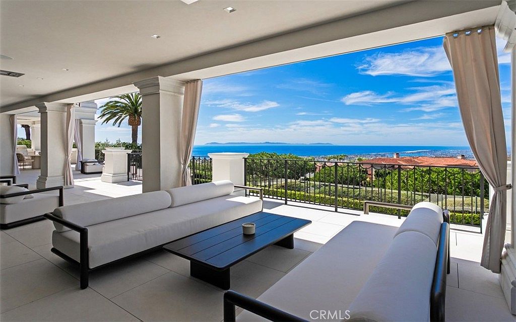 This expansive, covered outdoor patio offers a luxurious living space with a panoramic, unobstructed view of the ocean and distant coastline. The area is furnished with modern, minimalist white sofas and a sleek dark coffee table, creating an elegant lounge atmosphere perfect for entertaining. The open-air design, framed by classic columns and soft curtains, seamlessly blends indoor comfort with the breathtaking natural scenery.