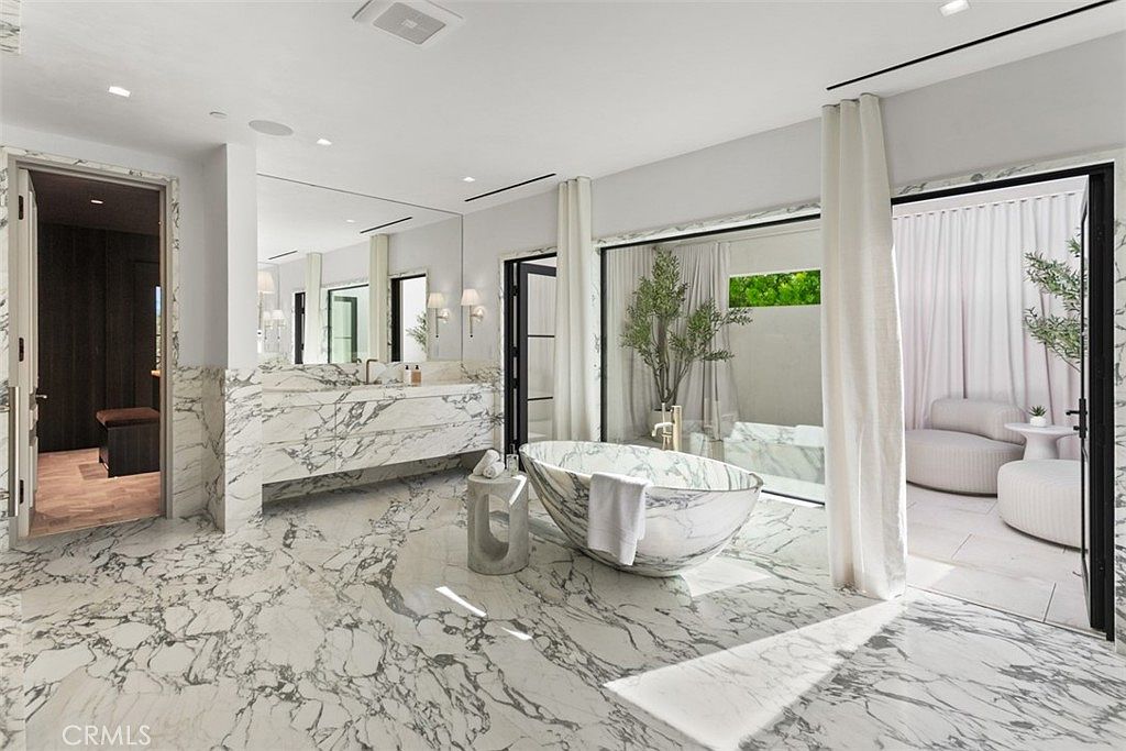 This luxurious primary bathroom features stunning floor-to-ceiling marble veining throughout, creating a sophisticated and high-end aesthetic. The space is anchored by a sculptural freestanding marble bathtub, complemented by a floating vanity and a seamless transition to an outdoor patio area through large glass doors. The bright, airy atmosphere is enhanced by soft drapery and minimalist design elements, offering a serene spa-like retreat.