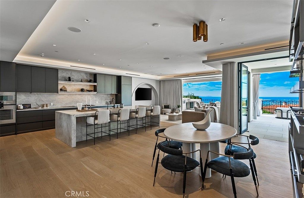 This expansive, open-concept kitchen and dining area showcases a sophisticated modern aesthetic with light hardwood flooring and a massive stone-topped island. The space flows seamlessly into a bright living area and an outdoor balcony that offers stunning ocean views, emphasizing a luxurious indoor-outdoor lifestyle. The design features sleek, dark cabinetry, minimalist bar seating, and a contemporary dining set, creating an atmosphere of refined elegance.