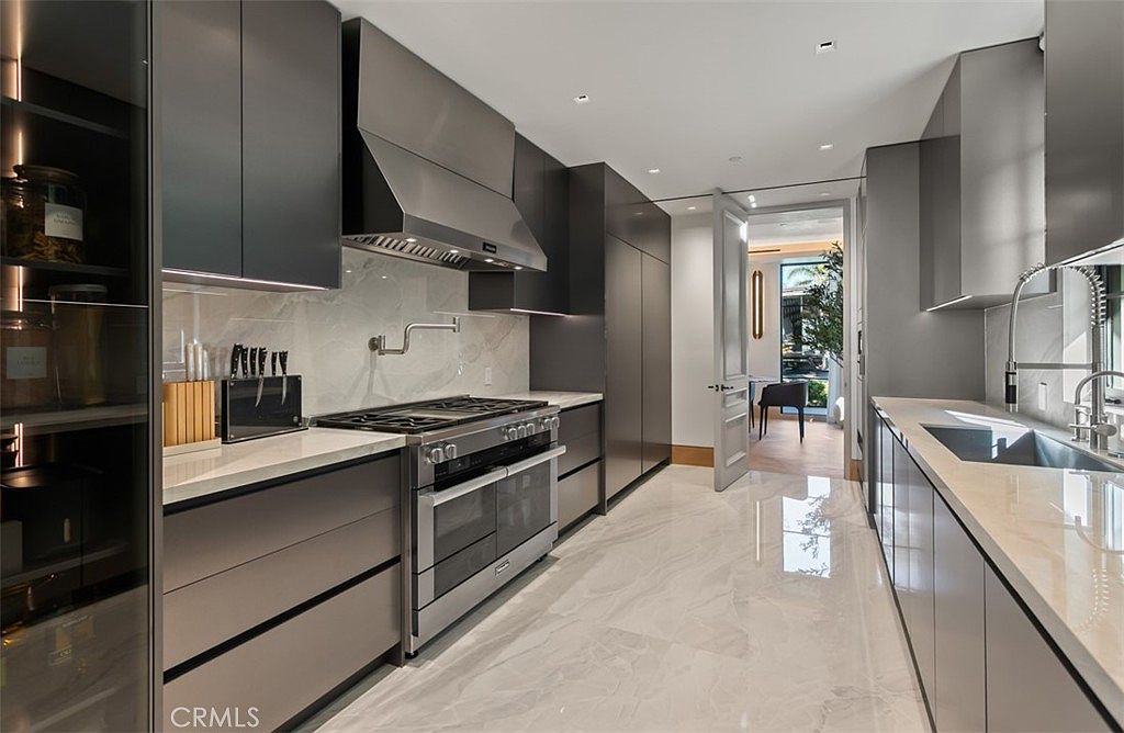 This sleek, modern kitchen features high-end dark grey cabinetry, polished marble-style flooring, and premium stainless steel appliances, including a professional-grade range. The space is illuminated by recessed lighting and under-cabinet accents, creating a sophisticated and minimalist aesthetic. A doorway leads into an adjacent dining area, providing a seamless flow between living spaces.