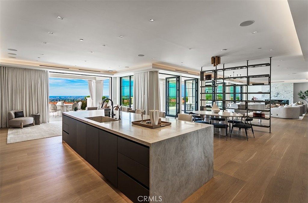 This expansive, open-concept kitchen features a massive stone-topped island with a built-in sink, serving as the centerpiece of a luxurious, modern living space. The room is bathed in natural light from floor-to-ceiling glass doors that open to a scenic outdoor view, while a sleek metal shelving unit separates the kitchen from the adjacent dining and living areas. The design aesthetic is sophisticated and airy, characterized by light wood flooring, recessed lighting, and a seamless transition between indoor and outdoor living.