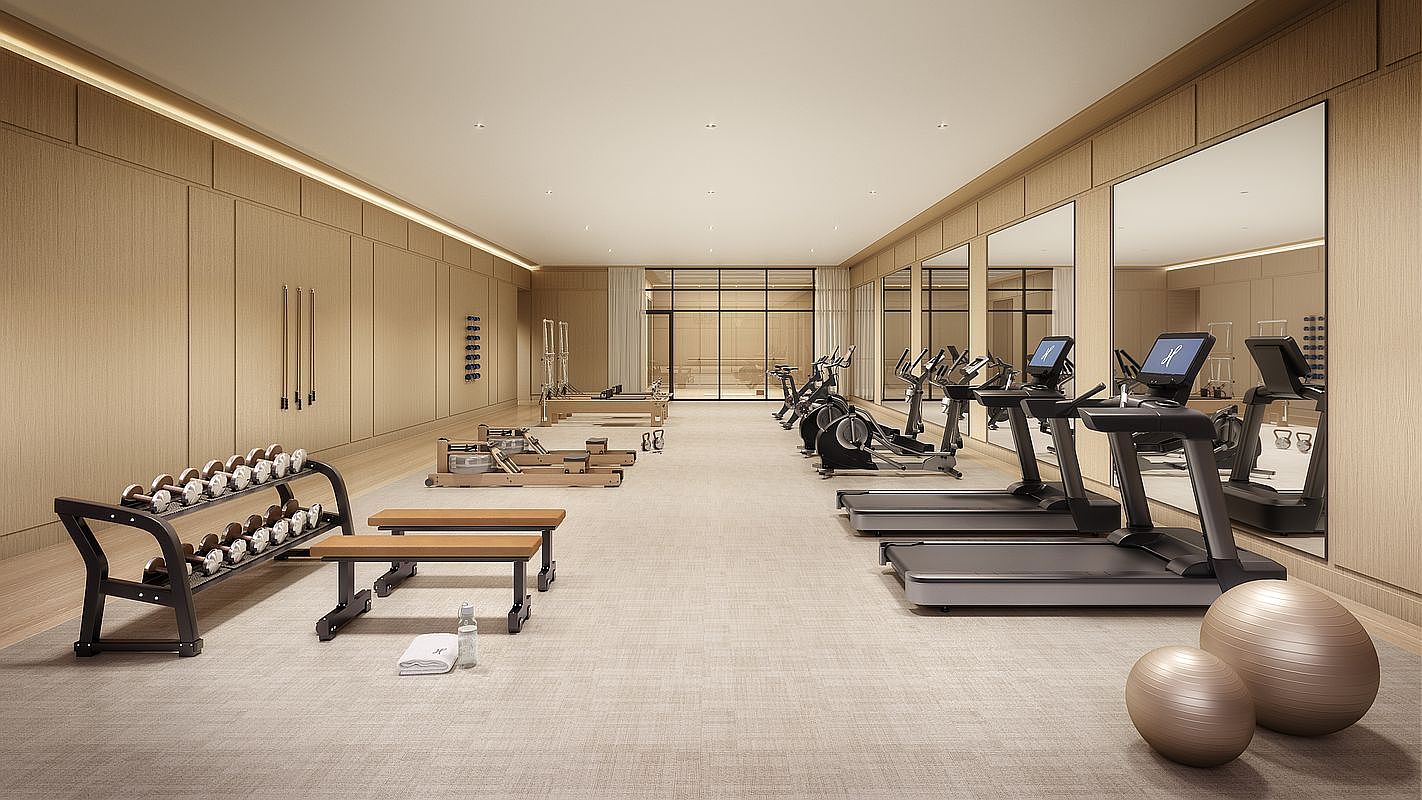 This spacious, modern home gym features light wood-paneled walls and a neutral carpeted floor, creating a serene and professional workout environment. The room is well-equipped with a rack of dumbbells, two benches, pilates reformers, stationary bikes, and treadmills positioned in front of large mirrors. The perspective is a wide-angle shot from the entrance, showcasing the clean, organized layout and the bright, airy atmosphere.