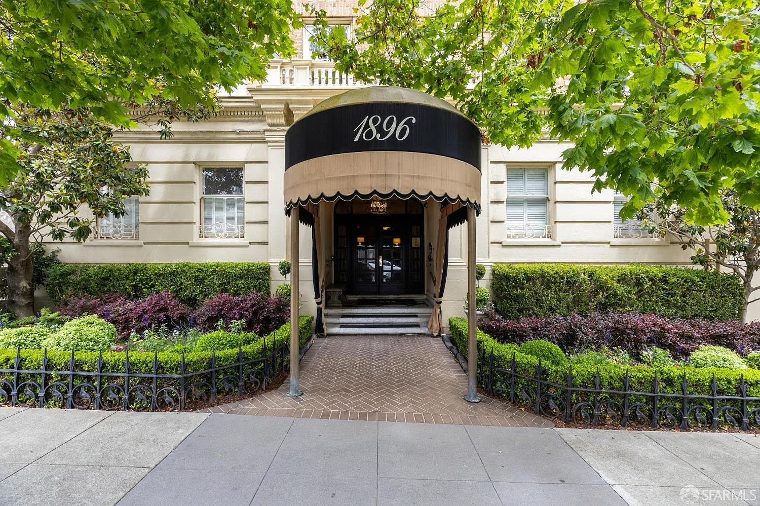 The image showcases the grand entryway of a building, featuring a prominent awning with the number "1896" displayed on top. The entrance is framed by manicured hedges and a wrought-iron fence, leading to a brick pathway. The building's facade is a light stone, exuding a sense of classic elegance and established prestige.