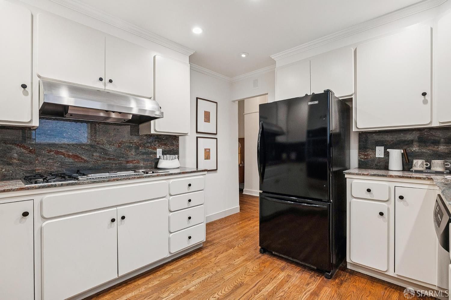 This is a well-lit kitchen featuring white cabinets with black hardware, granite countertops, and stainless steel appliances. A black refrigerator stands prominently against the wall, and hardwood floors add warmth to the space. The kitchen has a classic and functional design.