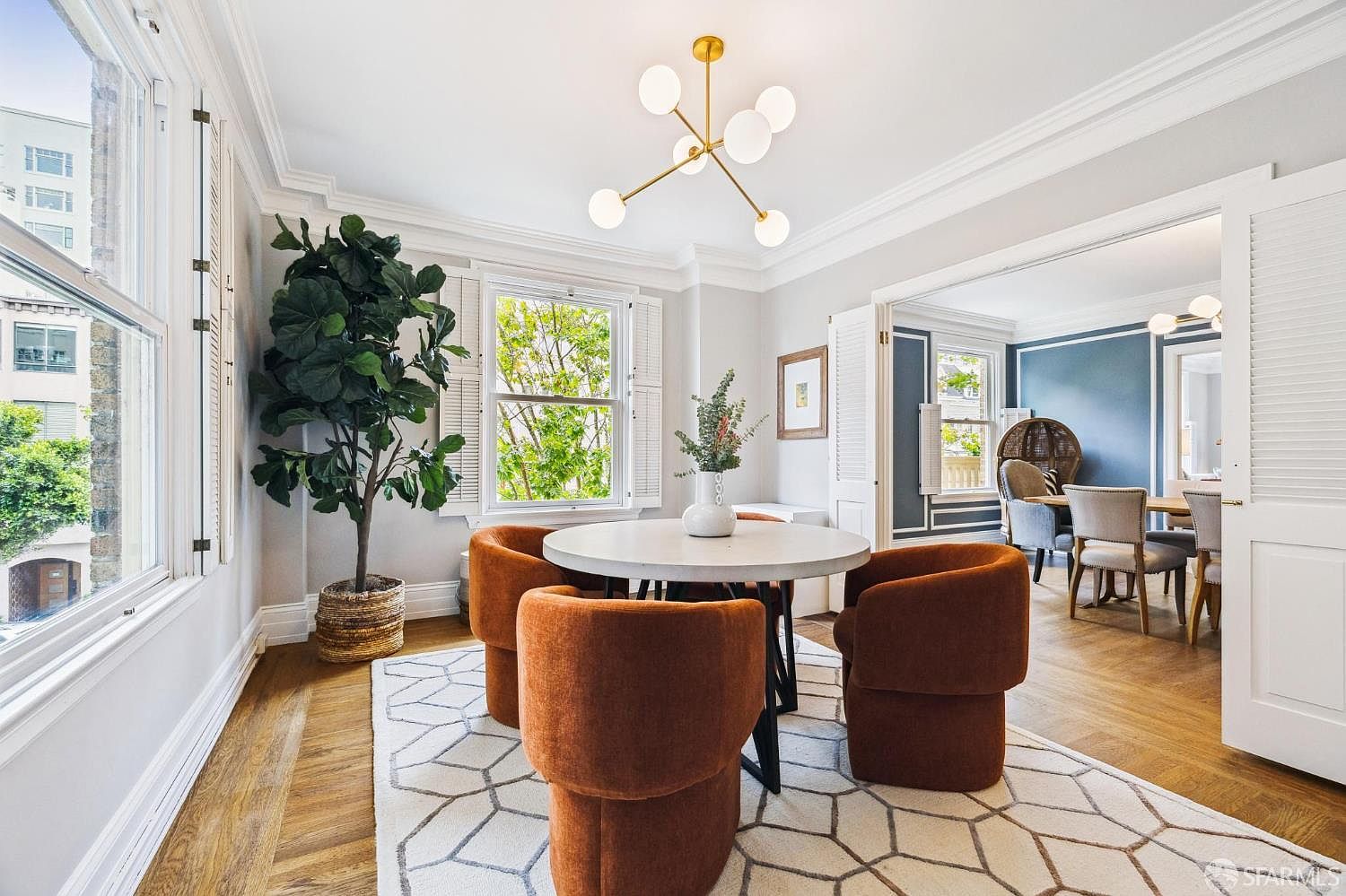 This is an interior shot of a dining room featuring a round table with four orange upholstered chairs. A modern chandelier hangs above the table, and a large potted plant sits near a window, adding a touch of nature to the space. The room is bright and airy, with hardwood floors and a stylish geometric rug, creating an inviting and sophisticated atmosphere.