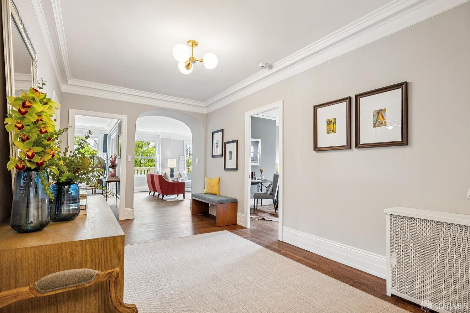 This interior shot showcases a well-lit hallway with hardwood floors and neutral-toned walls. The space is decorated with framed artwork and features an archway leading to a living area with red armchairs. A radiator cover and a console table with floral arrangements add to the home's charm and character.
