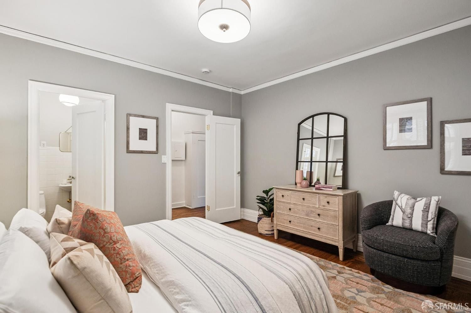 This is a well-lit primary bedroom featuring a neutral color palette with gray walls and white trim. The room includes a bed with decorative pillows, a wooden dresser with a mirror, and a cozy armchair. Artwork adorns the walls, and the hardwood floors add warmth to the space, creating a serene and inviting atmosphere.