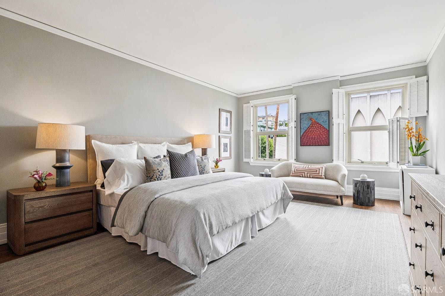 This is a bright and airy primary bedroom featuring a large bed with neutral-toned bedding and decorative pillows. The room is painted in a soft gray, complemented by white trim and window shutters. Natural light floods the space through multiple windows, highlighting the hardwood floors and a comfortable chaise lounge.