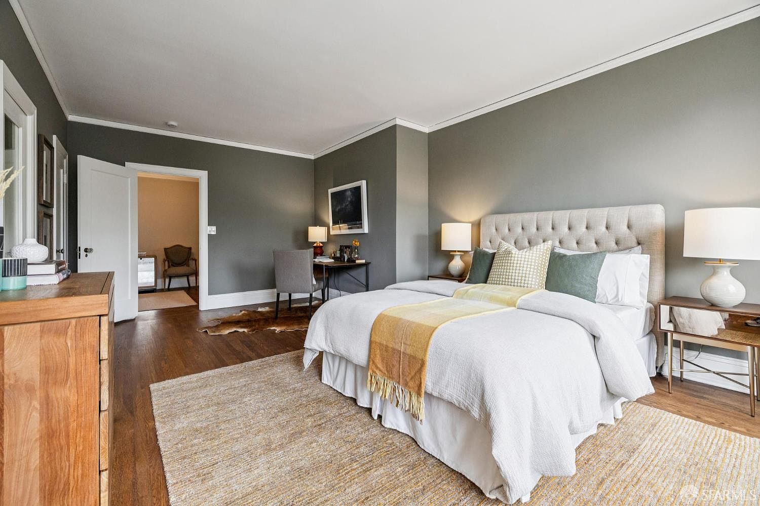 This is a primary bedroom featuring a neutral color palette with gray walls and white trim. The room includes a large bed with a tufted headboard, a side table with a lamp, and a desk area with a chair. A doorway leads to another room, and a rug covers the hardwood floor, creating a cozy and inviting atmosphere.