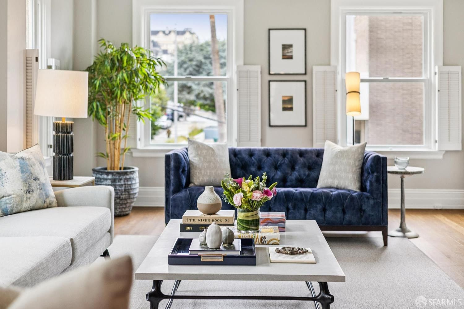 This is a beautifully staged living room featuring a plush blue velvet sofa with tufted detailing, complemented by neutral-toned pillows and a light-colored area rug. A modern coffee table displays decorative items and books, while natural light streams in through the windows, enhancing the room's inviting atmosphere. The perspective is from a medium shot, showcasing the room's depth and arrangement.