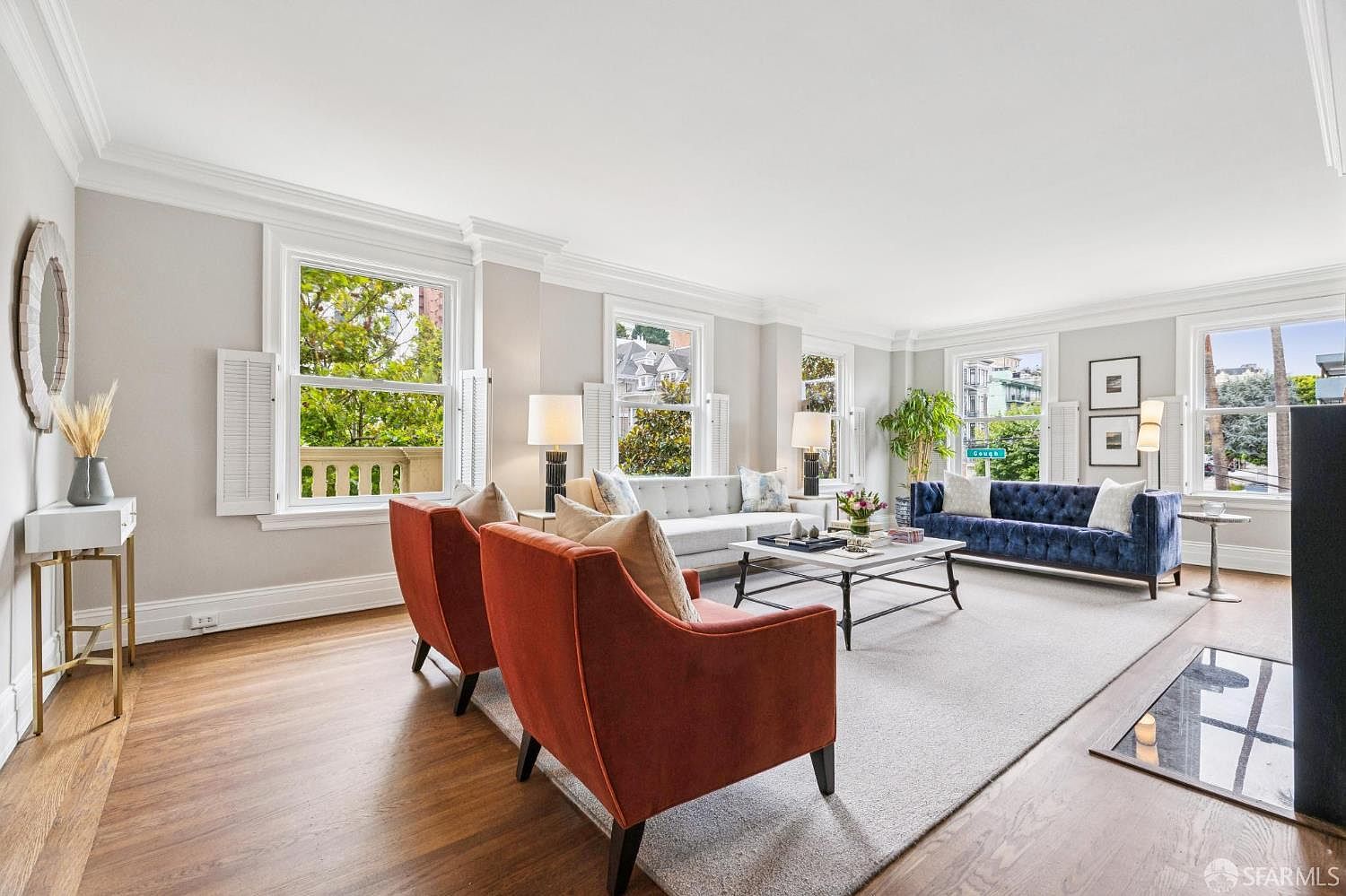 This is a spacious living room featuring hardwood floors, a neutral color palette, and ample natural light from large windows. The room is furnished with two orange armchairs, a light gray sofa, a blue velvet sofa, a marble coffee table, and various decorative items. The overall impression is elegant and inviting, perfect for entertaining or relaxing.