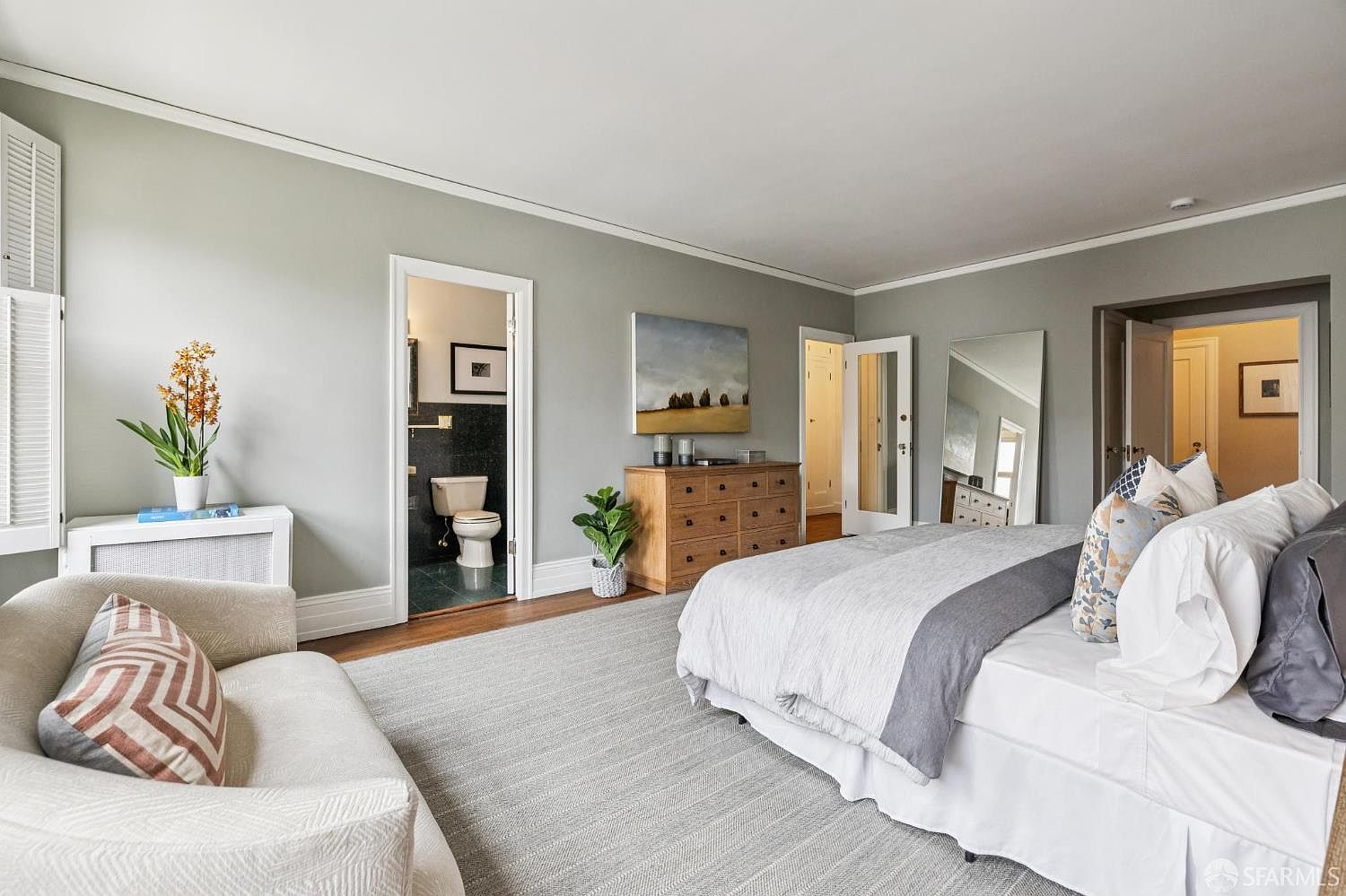This is a spacious primary bedroom featuring a large bed with white linens and a gray throw, complemented by decorative pillows. The room is painted in a neutral gray tone, with white trim and hardwood floors partially covered by a light gray rug. There is a dresser with artwork above it, a comfortable armchair, and doorways leading to other rooms, including one to a bathroom.