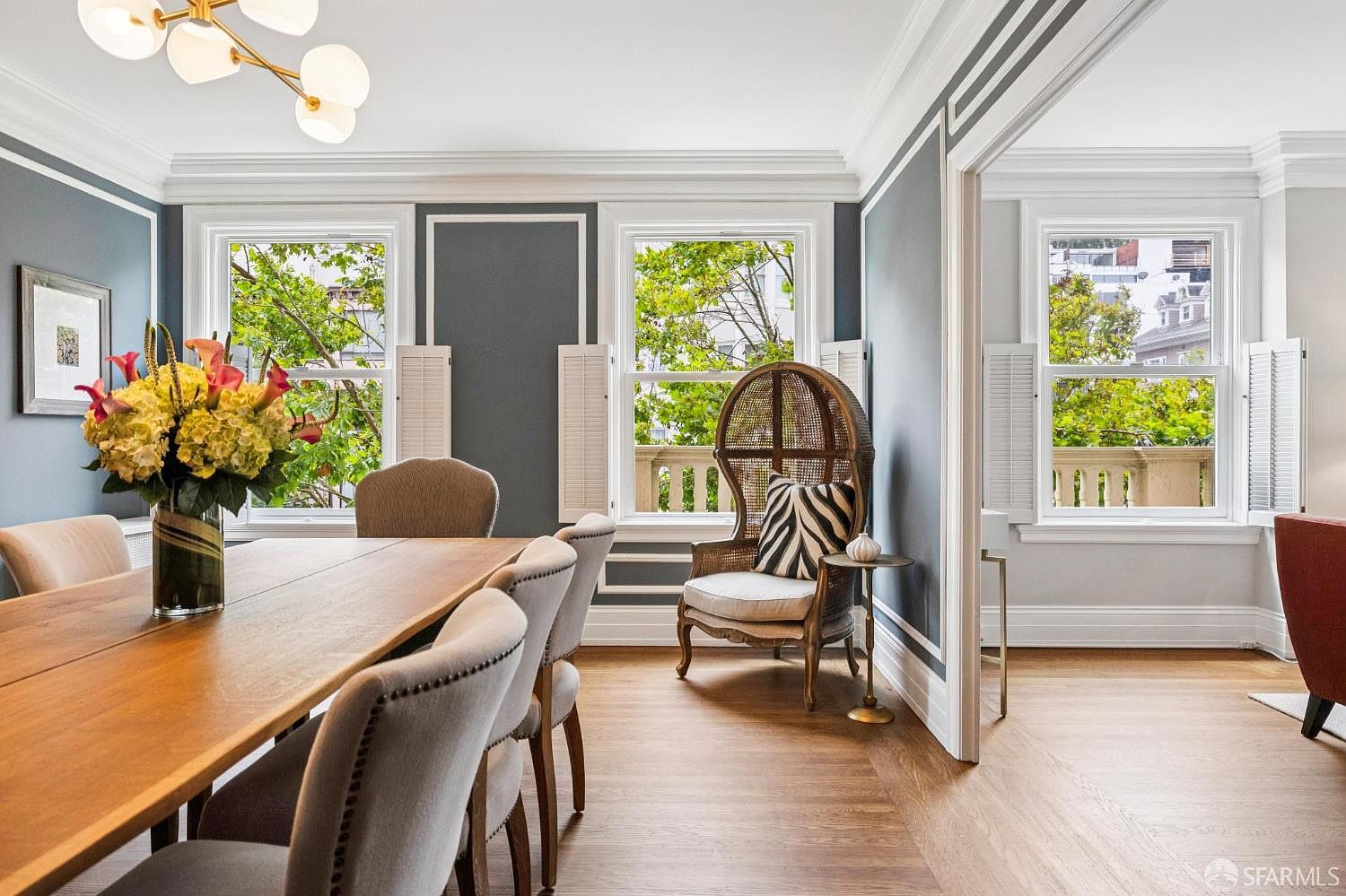 This is a dining room featuring a long wooden table with upholstered chairs. A vase of flowers sits on the table, adding a touch of color. The room has a sophisticated feel with gray walls, white trim, and large windows that provide natural light and views of greenery outside. A decorative peacock chair sits near the window.