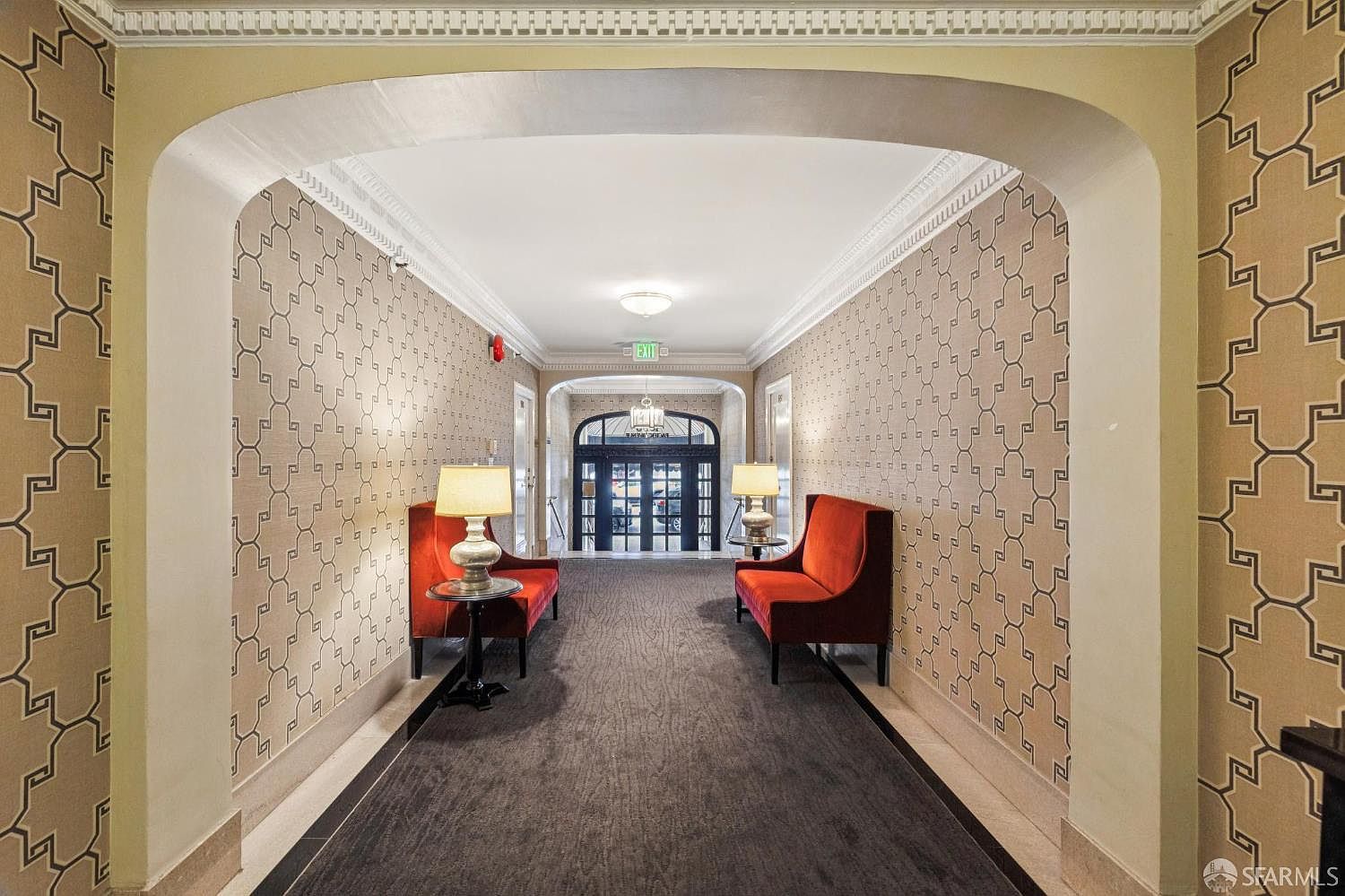This interior shot showcases a well-appointed hallway with patterned wallpaper and elegant archways. Two red settees with lamps on side tables flank the hallway, creating a welcoming and sophisticated ambiance. The dark carpet runner and marble flooring add to the luxurious feel, leading towards a set of glass doors at the end of the hall.