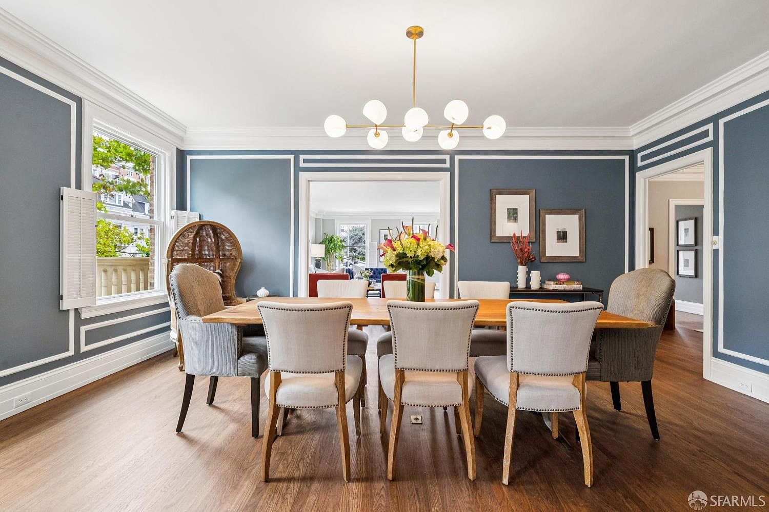 This is an interior shot of a dining room featuring a long wooden table surrounded by upholstered chairs with wooden legs. A modern chandelier hangs above the table, and artwork adorns the blue walls. The room has hardwood floors and white trim, creating a sophisticated and inviting atmosphere.