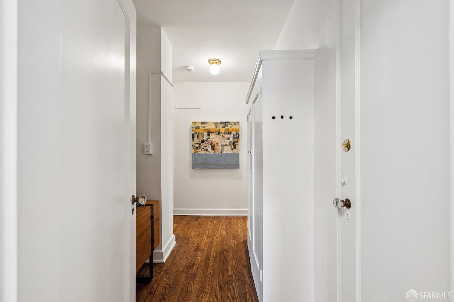 This is an interior shot of a hallway featuring hardwood floors and white walls. An abstract painting hangs at the end of the hallway, illuminated by a ceiling light. The perspective gives a sense of depth and leads the eye down the corridor, showcasing the clean lines and simple design.