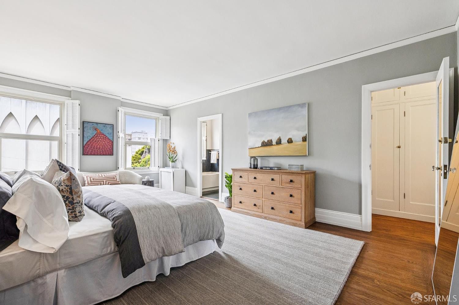 This is a primary bedroom featuring a large bed with gray and white bedding, a wooden dresser with a landscape painting above it, and two windows with white shutters. The walls are painted a light gray, and the floors are hardwood with a large area rug. The room has a calm and inviting atmosphere.