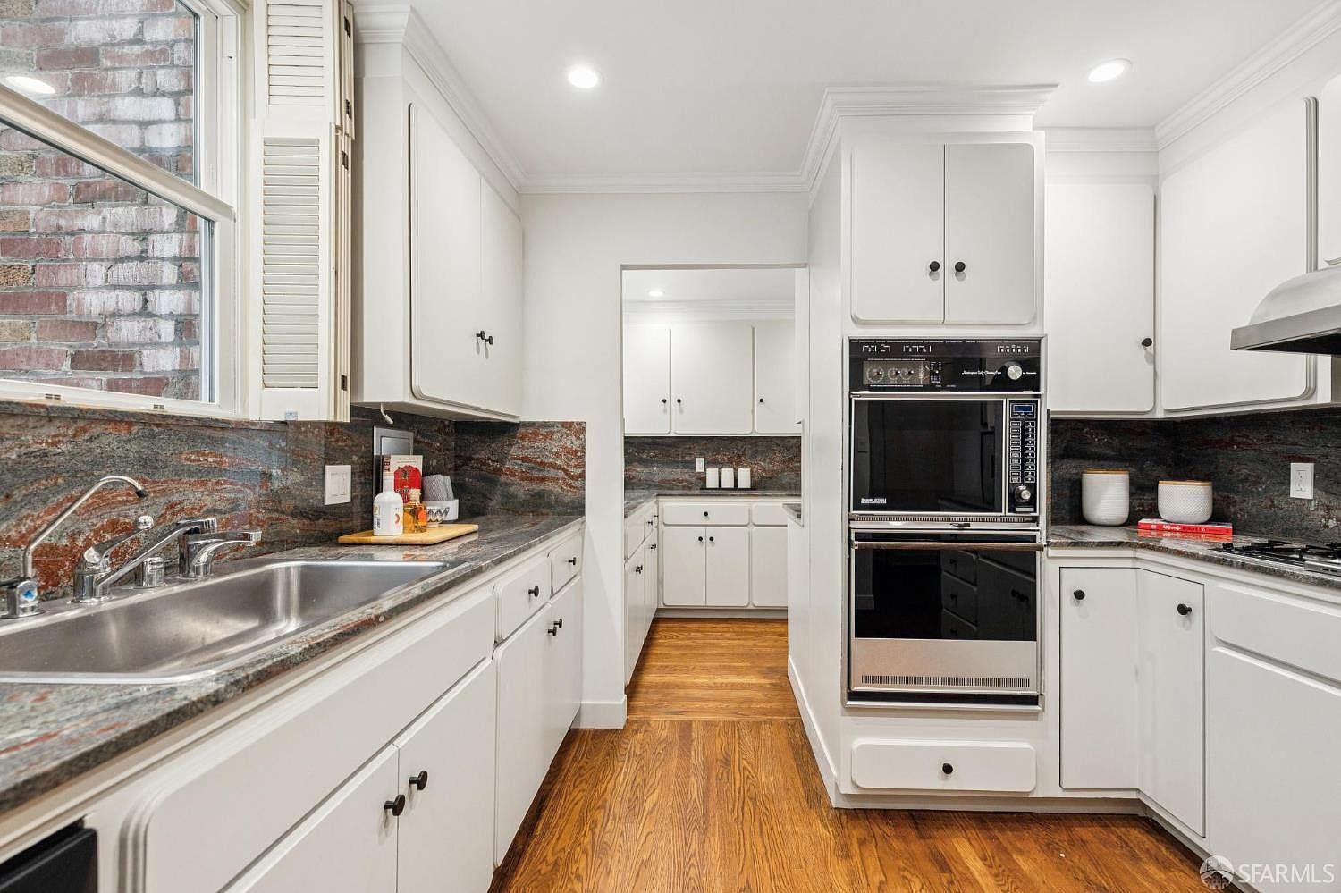 This is a well-lit kitchen featuring white cabinetry, dark countertops with reddish-brown veining, and stainless steel appliances. The kitchen has a classic design with a built-in oven and microwave, and hardwood flooring adds warmth to the space. A window provides natural light, and the overall impression is clean and functional.