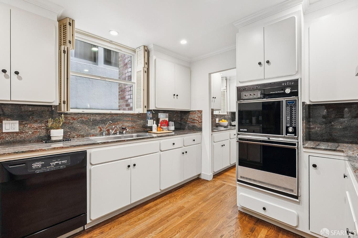 This is a well-lit kitchen featuring white cabinetry with black hardware, a black dishwasher, and a built-in double oven. The countertops are a dark, speckled stone, and the flooring is hardwood. A window above the sink provides natural light, and the overall impression is a clean and functional space.