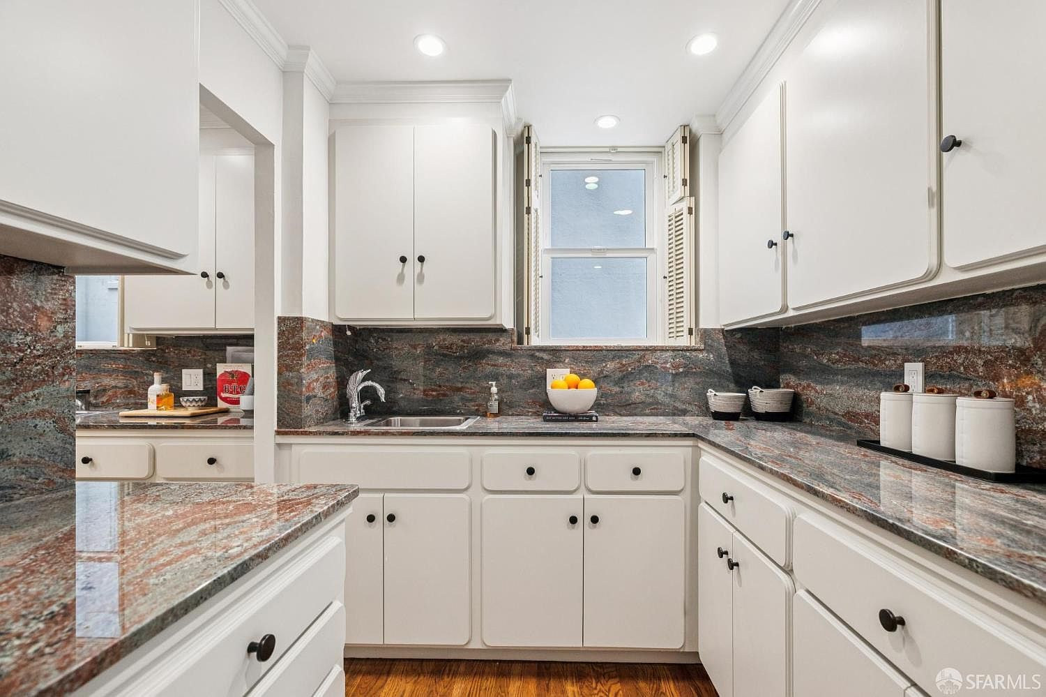 This is a well-lit kitchen featuring white cabinetry with dark hardware and a unique granite countertop and backsplash. A window above the sink provides natural light, and the hardwood flooring adds warmth to the space. The kitchen appears clean and functional, with a classic design.