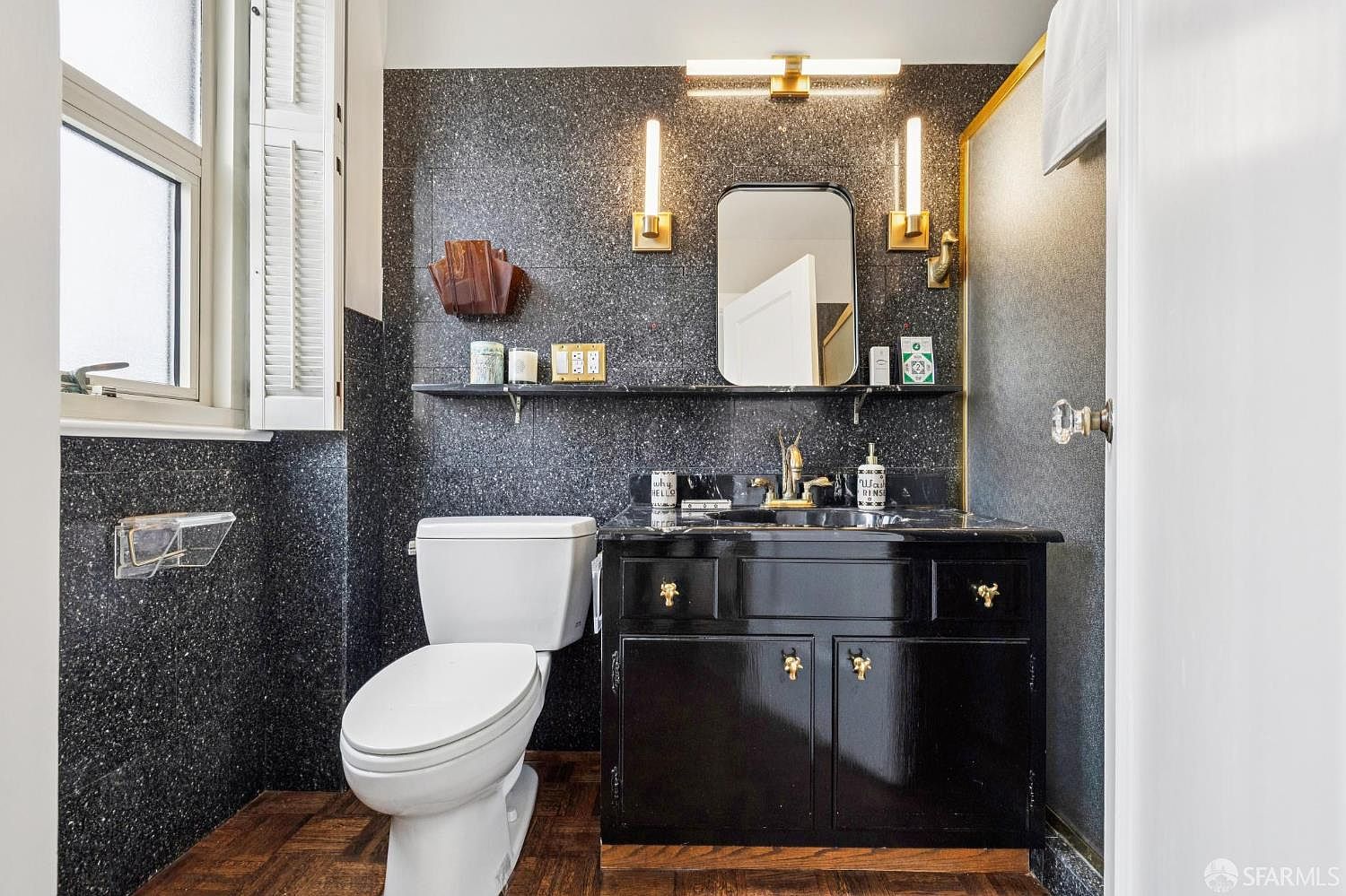 This is a guest bathroom featuring a black speckled wall design, a black vanity with gold hardware, and a white toilet. A shelf above the toilet holds decorative items, and the lighting fixtures above the mirror add a touch of elegance. The overall impression is a stylish and well-maintained space.